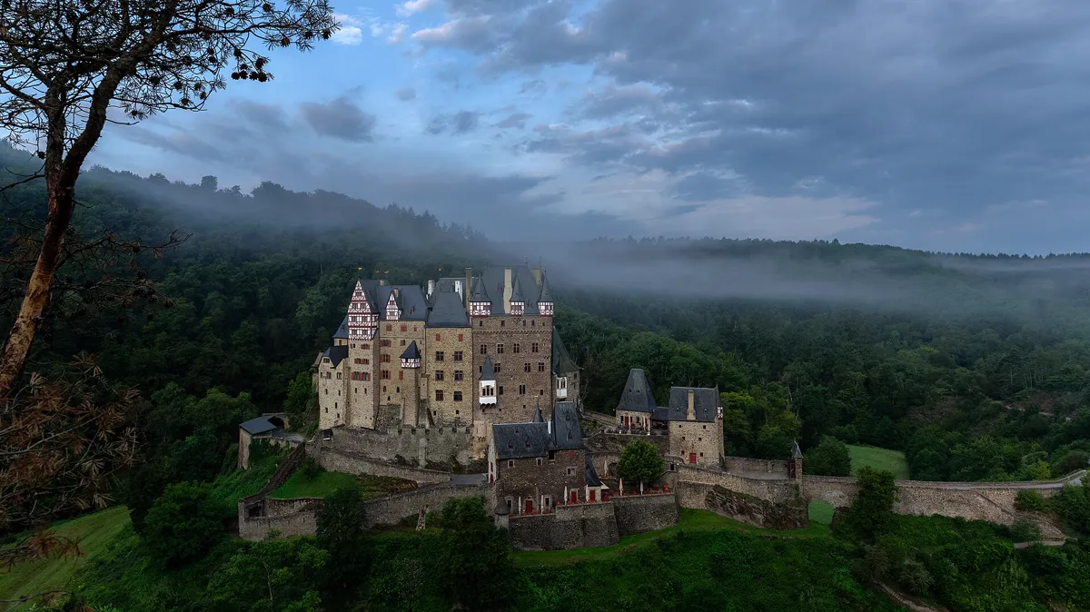 Hilltop Castle With Slate Turrets Above a Misty Forest Valley Stone castle with slate turrets above a misty forest valley in early light, a fitting scene for a throne of glass reading order guide